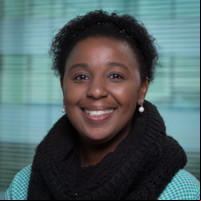 A foto mostra o rosto de uma mulher sorridente sobre um fundo verde-água. Ela é negra, tem cabelos curtos e escuros e olhos também escuros, e veste um cachecol preto sobre uma camisa verde-água.